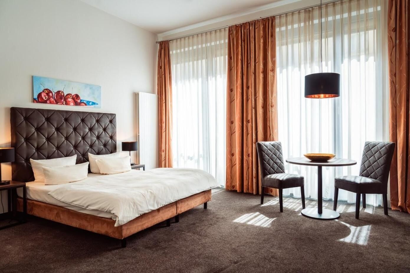 A hotel room with a large bed, a bedside table, a lamp, a painting, a round table with two chairs, and a large window with curtains.