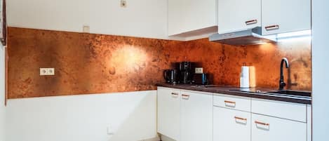 A compact kitchen with white cabinets, a copper backsplash, and a black faucet.
