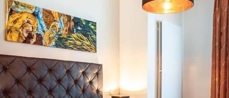 Une chambre d’hôtel avec une tête de lit capitonnée, un grand tableau, une lampe sur pied et un lit recouvert de draps blancs.