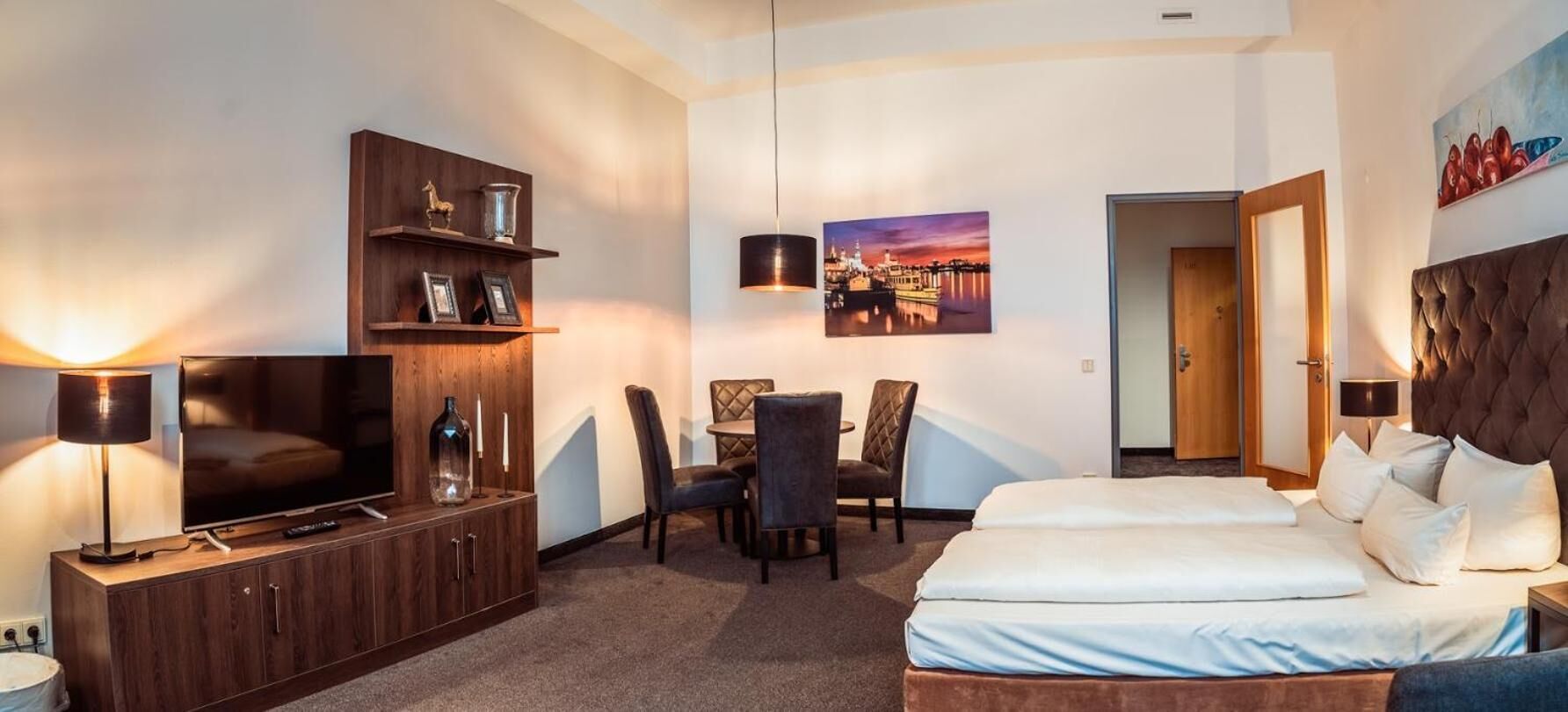 Une chambre d’hôtel équipée d’une télévision, d’une table à manger avec des chaises, d’un lit et d’un tableau au mur.
