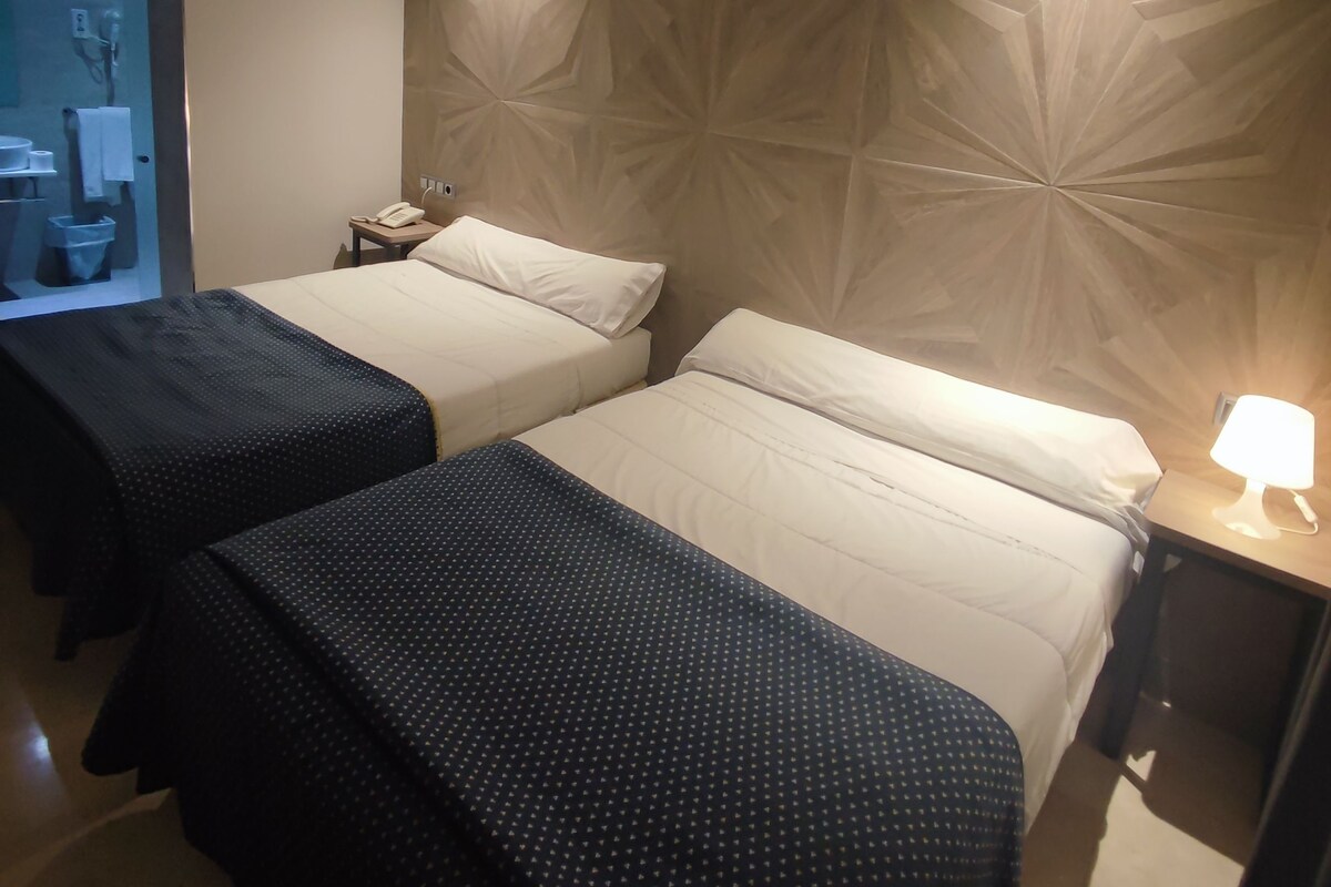 Habitación de hotel con dos camas, un cabecero de madera, una mesita de noche con lámpara, y un baño visible a través de una puerta de cristal.