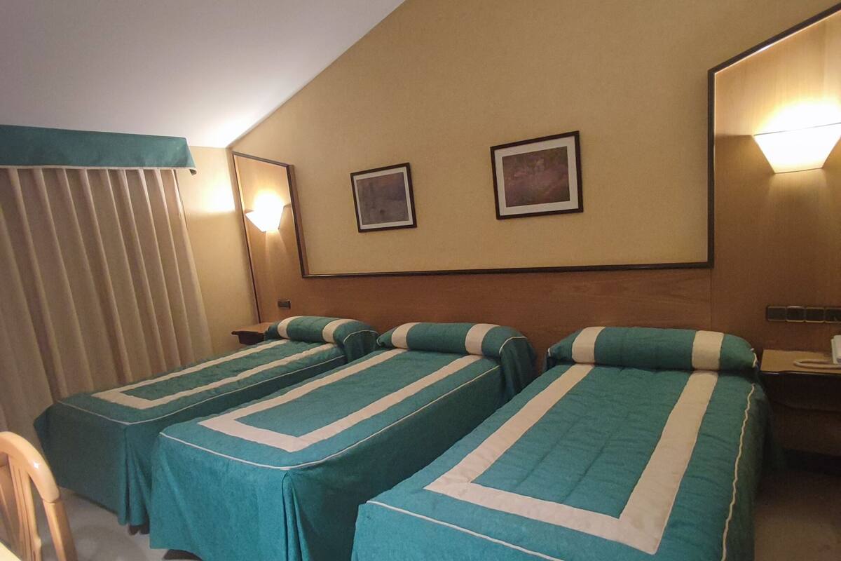 Habitación de hotel con dos camas individuales, un ventanal con persianas, un espejo y dos lámparas de pared.