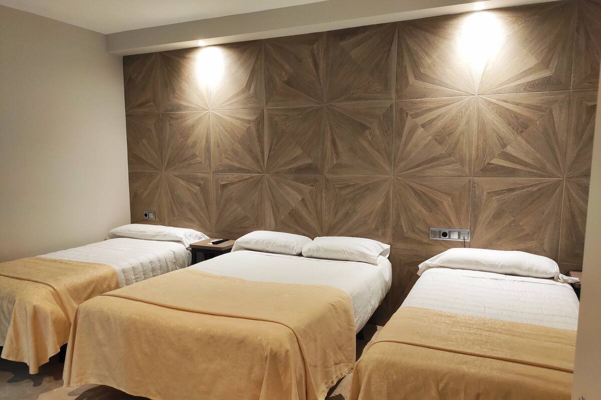 Habitación de hotel con tres camas, cada una con cubrecama color crema y almohadas blancas. El cabecero presenta un diseño geométrico de paneles de madera.