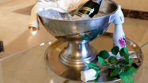 Un saladier en métal, une bouteille et des verres posés sur une table en verre, une rose blanche et une bouteille d’eau.
