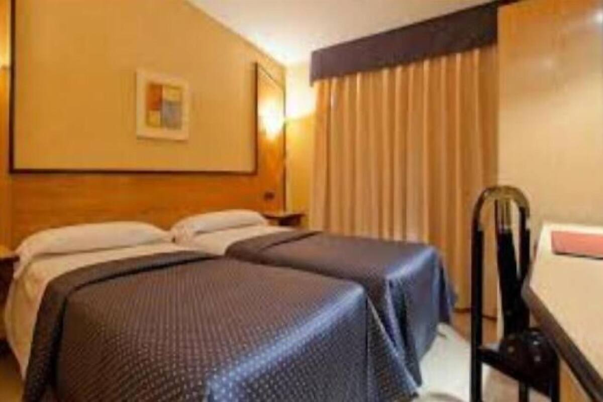Habitación de hotel con dos camas individuales, un cabecero de madera, una mesita de noche pequeña y una silla.