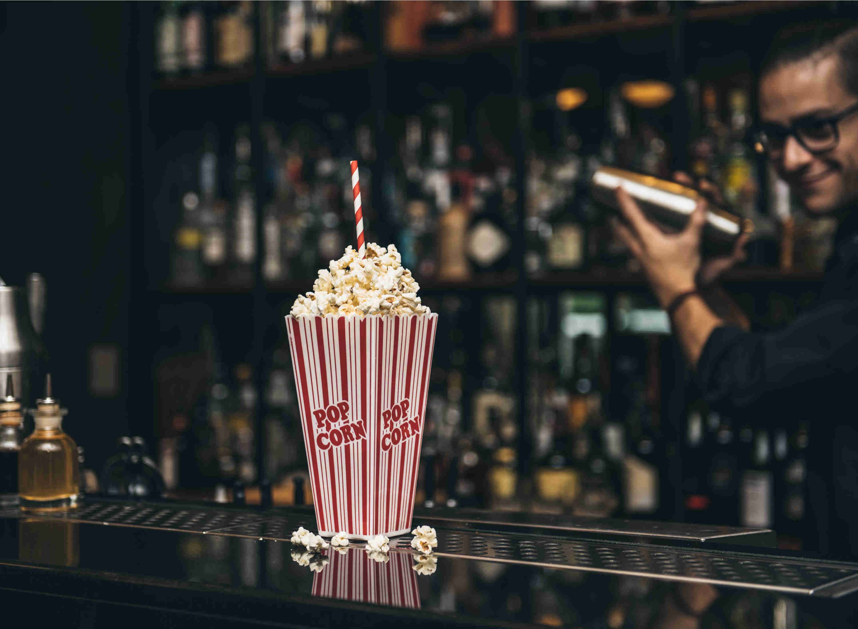 A bar with a striped popcorn container labeled "Pop Corn" and a person holding a drink.