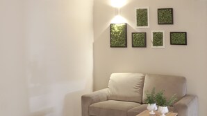 A living room with a beige sofa, a matching ottoman, a small wooden side table with two white vases, and a wall-mounted light.