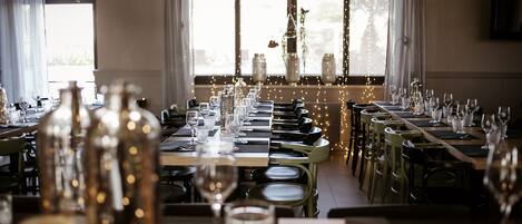 Een restaurant met lange tafels gedekt voor het dineren, grote ramen met gordijnen en sfeerverlichting.