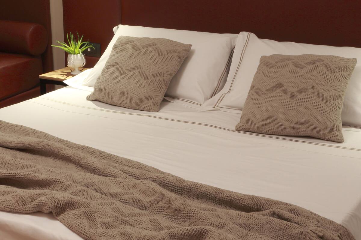 A neatly made bed with a beige blanket, two matching pillows, and a small potted plant on a bedside table.