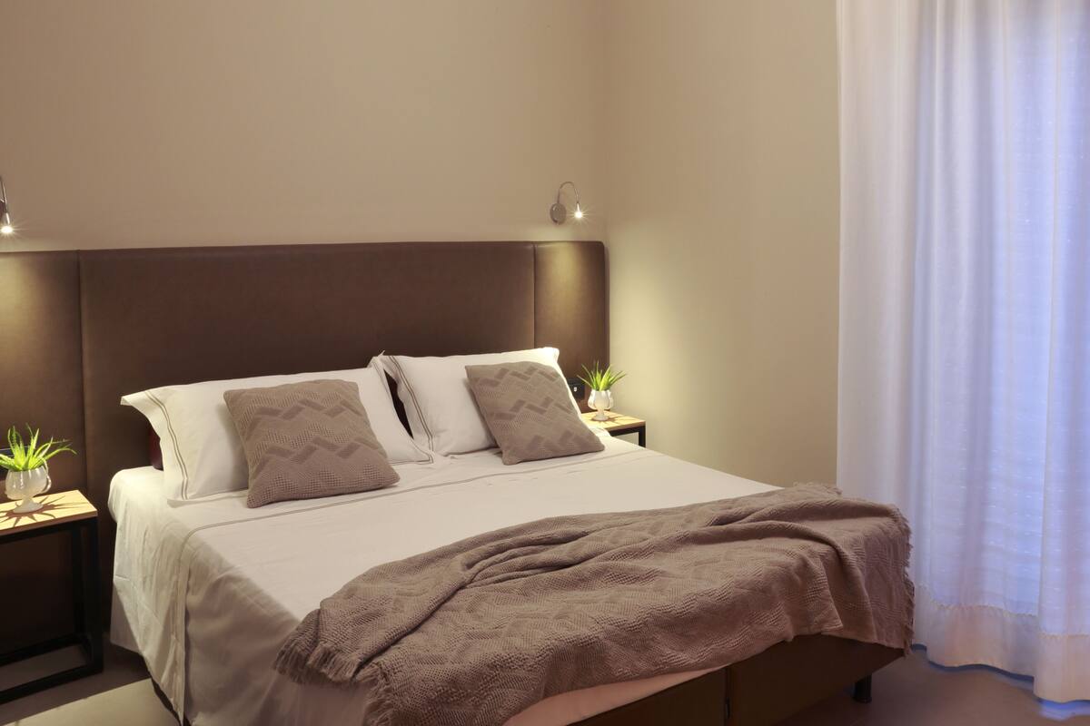A neatly made bed with a brown blanket, two bedside tables with lamps, and a window with white curtains.