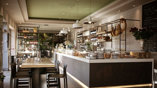 A modern restaurant interior with a central bar area, seating arrangements, and a variety of displayed items on shelves.