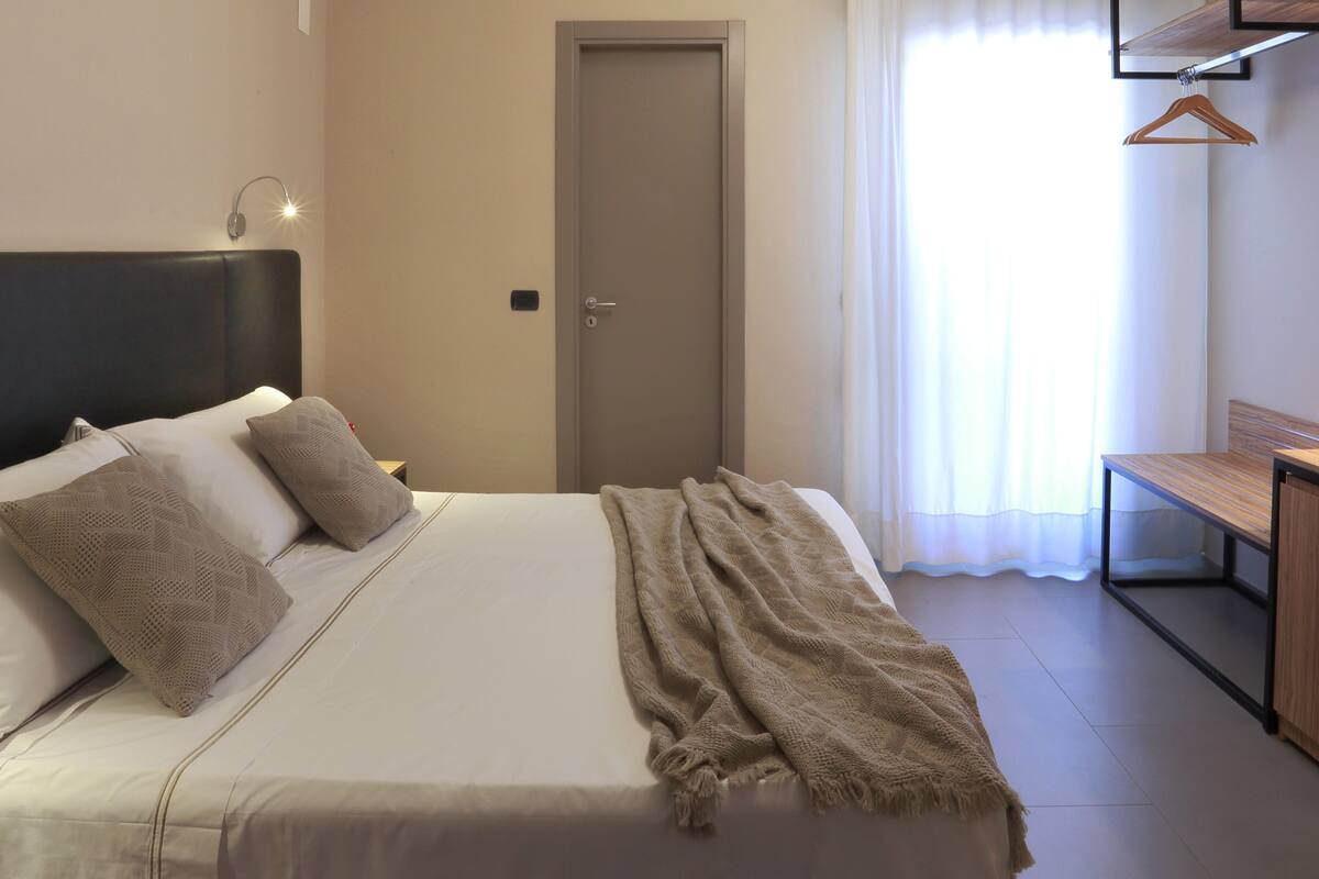 A neatly made bed with pillows and a blanket, a bedside table, a door, and a window with curtains.
