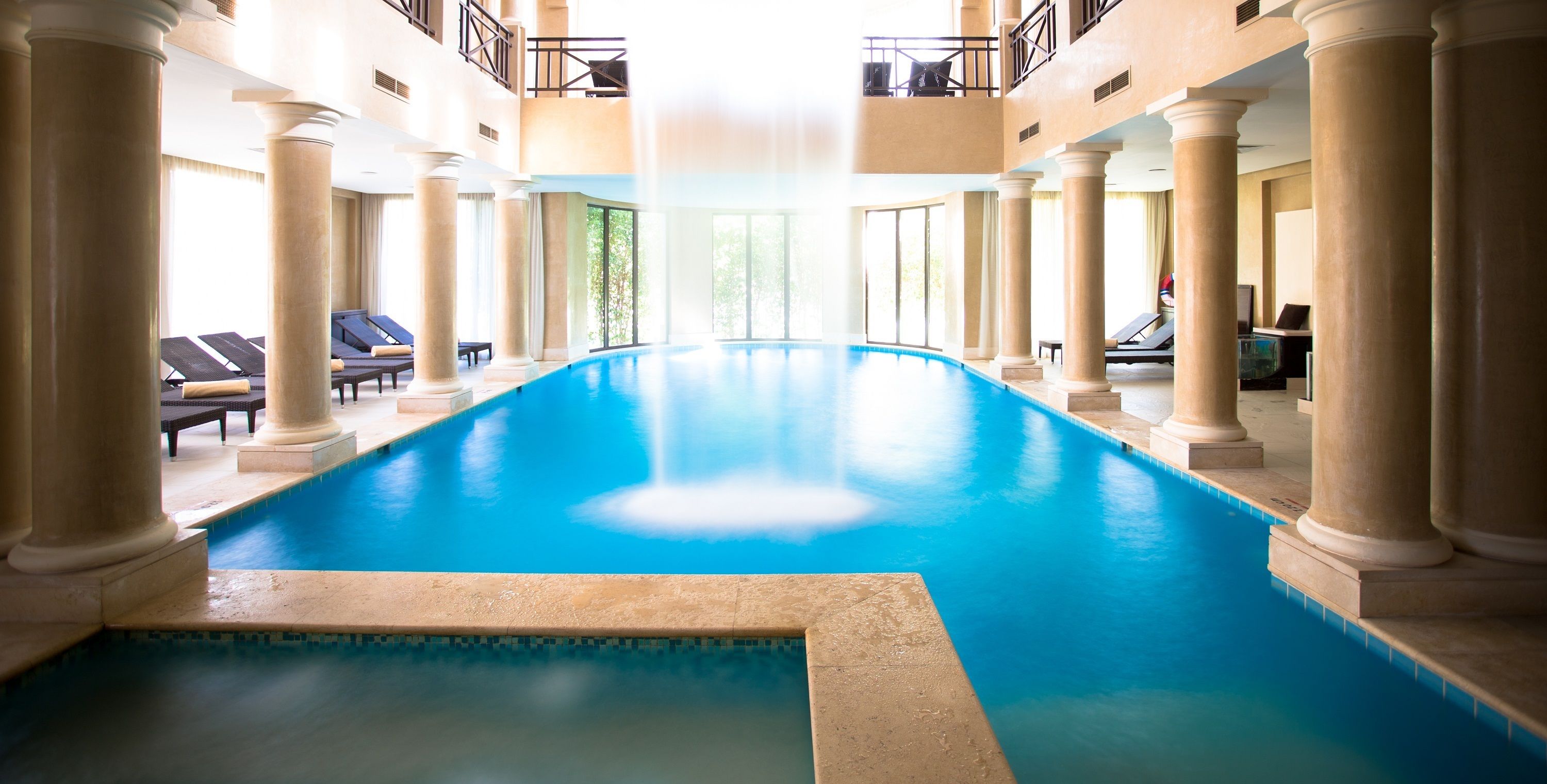 An indoor swimming pool with columns, sun loungers, and a view of the outdoors.