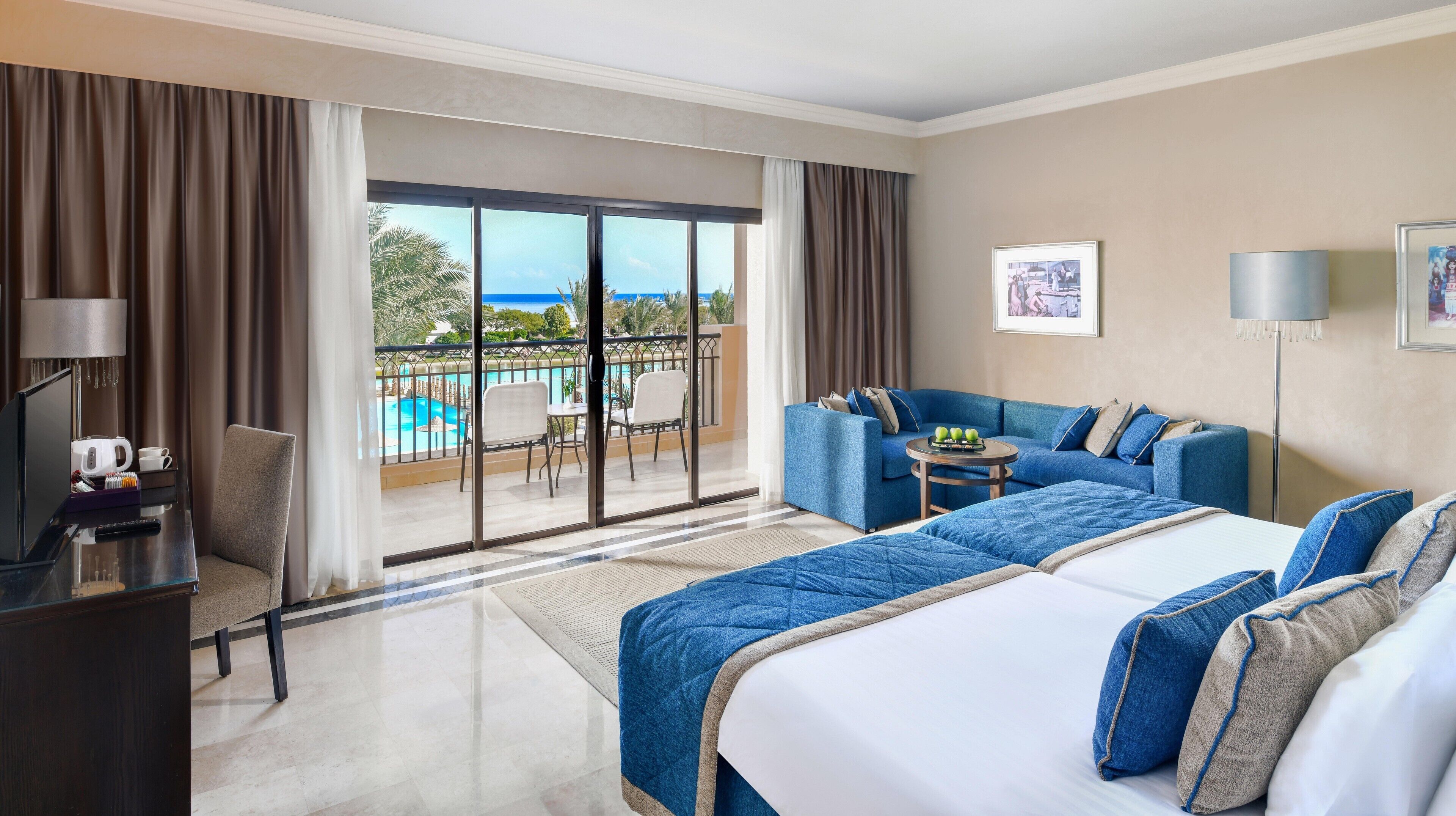 A hotel room with a large bed, a sofa, a desk with a lamp, and a view of a pool and palm trees.