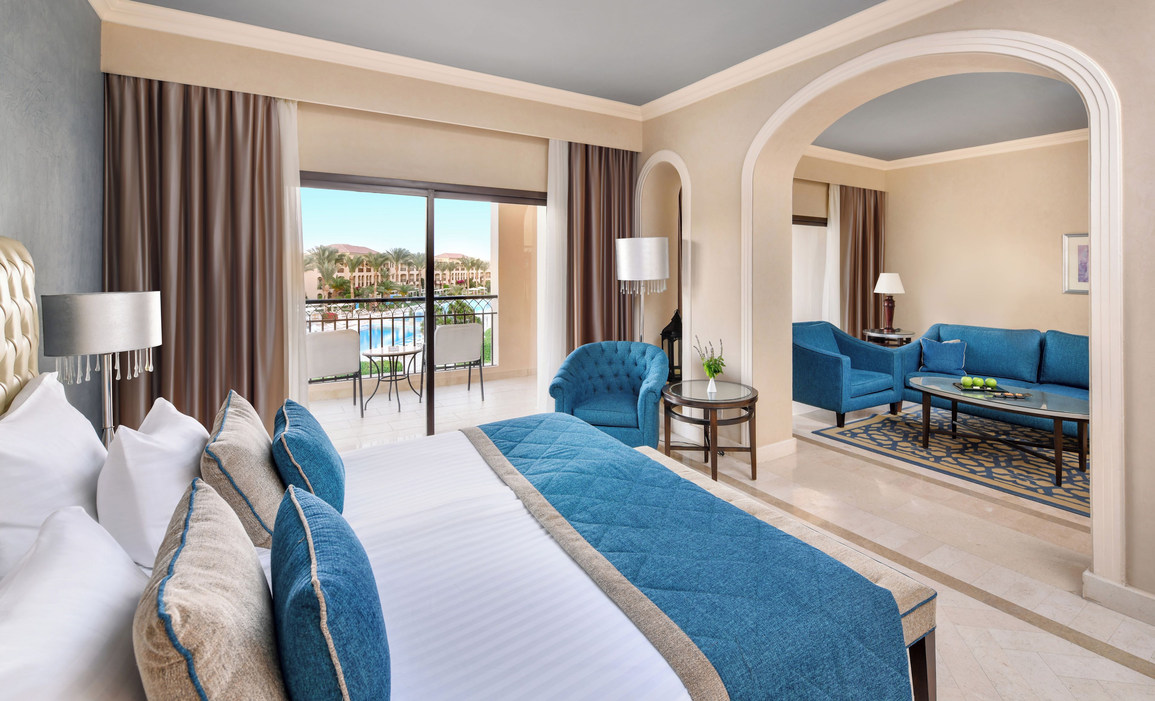 A hotel room with a bed, a sofa, a coffee table, and a view of a pool and buildings.