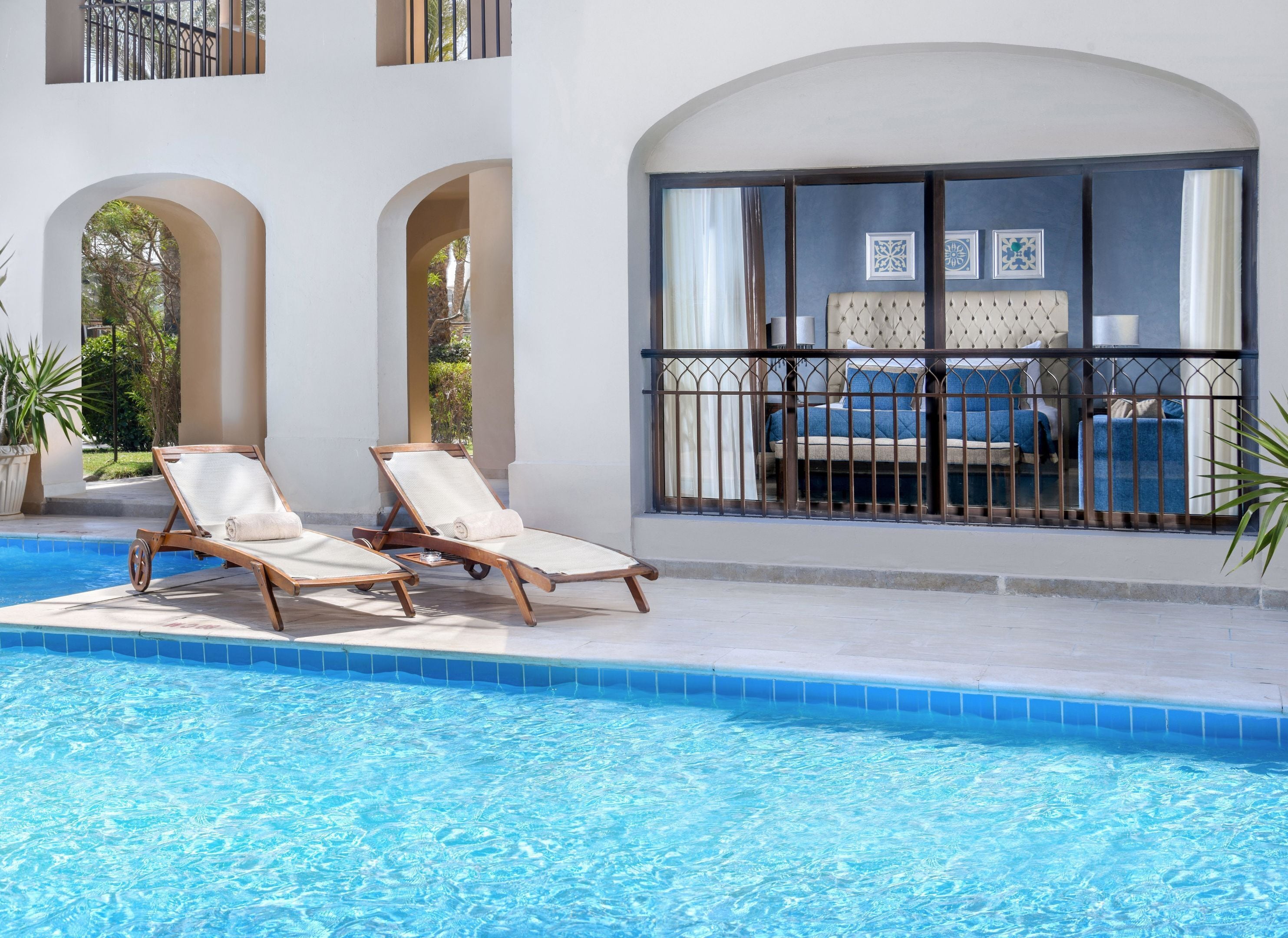 Un espace aménagé au bord de la piscine, avec deux chaises longues, une grande fenêtre ornée de cadres décoratifs et une vue sur un immeuble doté de balcons.