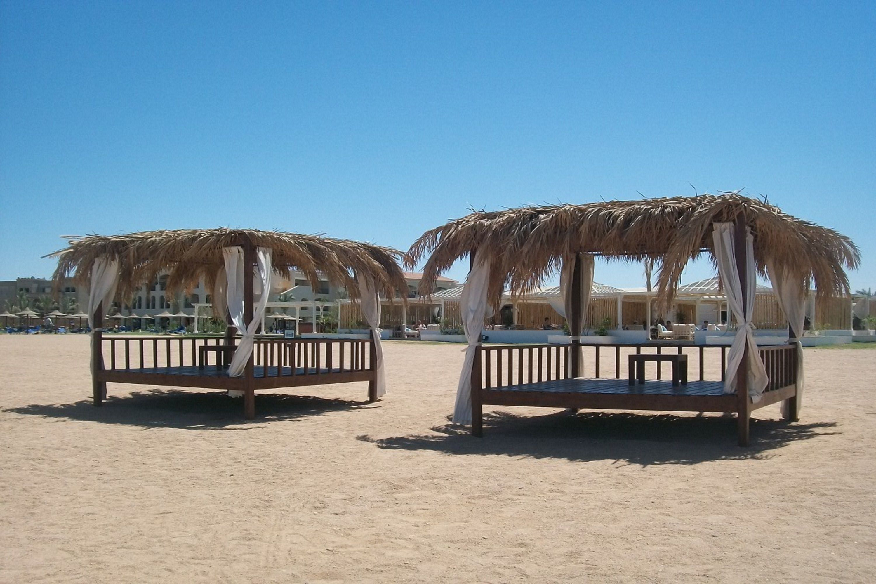 Two thatched-roof beds with white curtains, set on a sandy beach with buildings in the background.