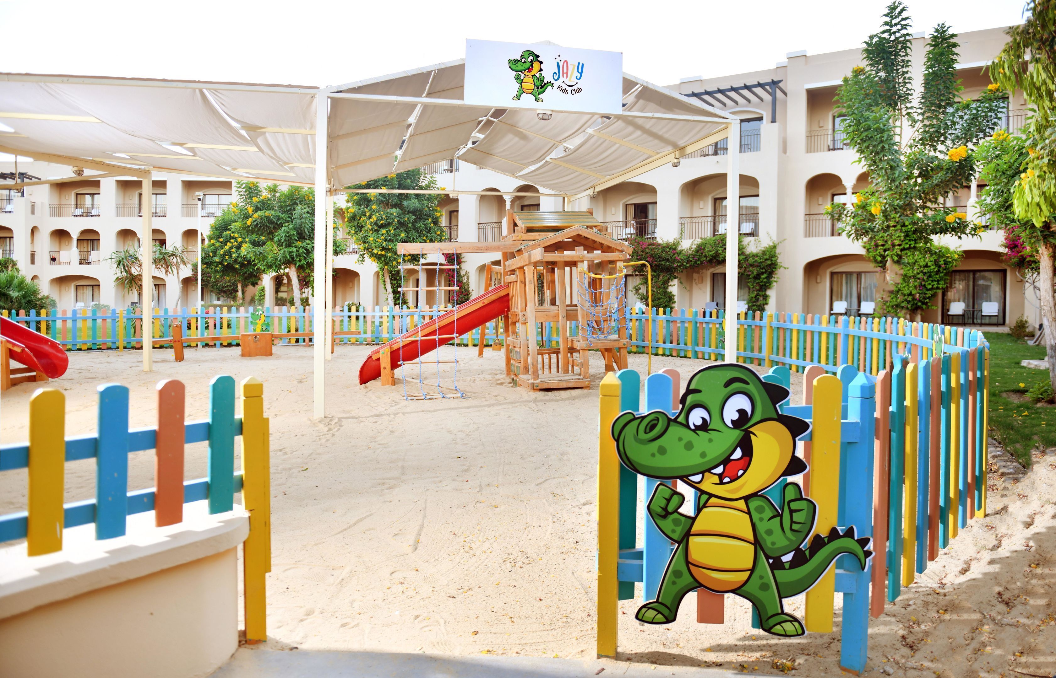Zona infantil con tobogán y estructuras para escalar, rodeada por una valla colorida y un edificio con balcones al fondo.