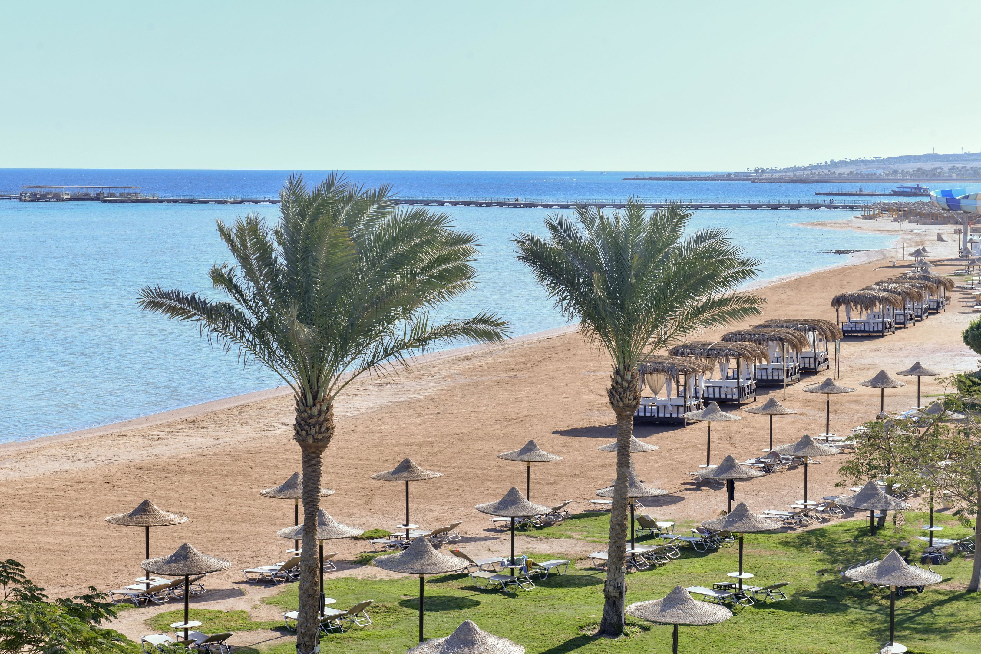 A beach resort with rows of sun loungers and thatched umbrellas, a clear view of the sea, and a distant pier.