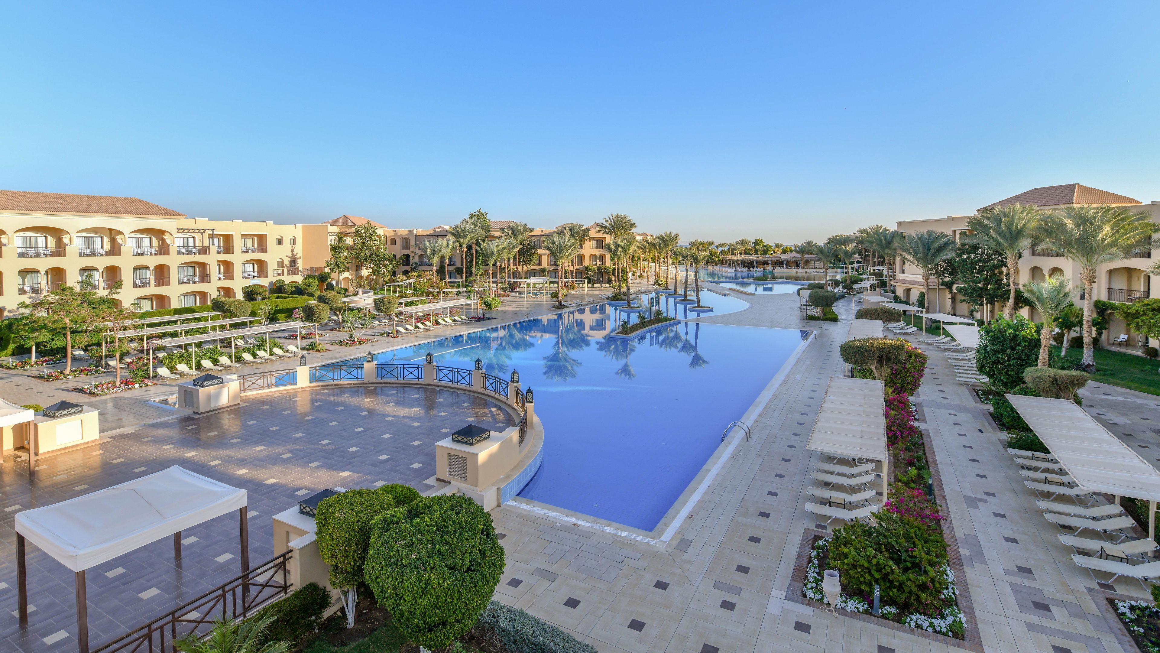 A resort with a large swimming pool, sun loungers, and palm trees.