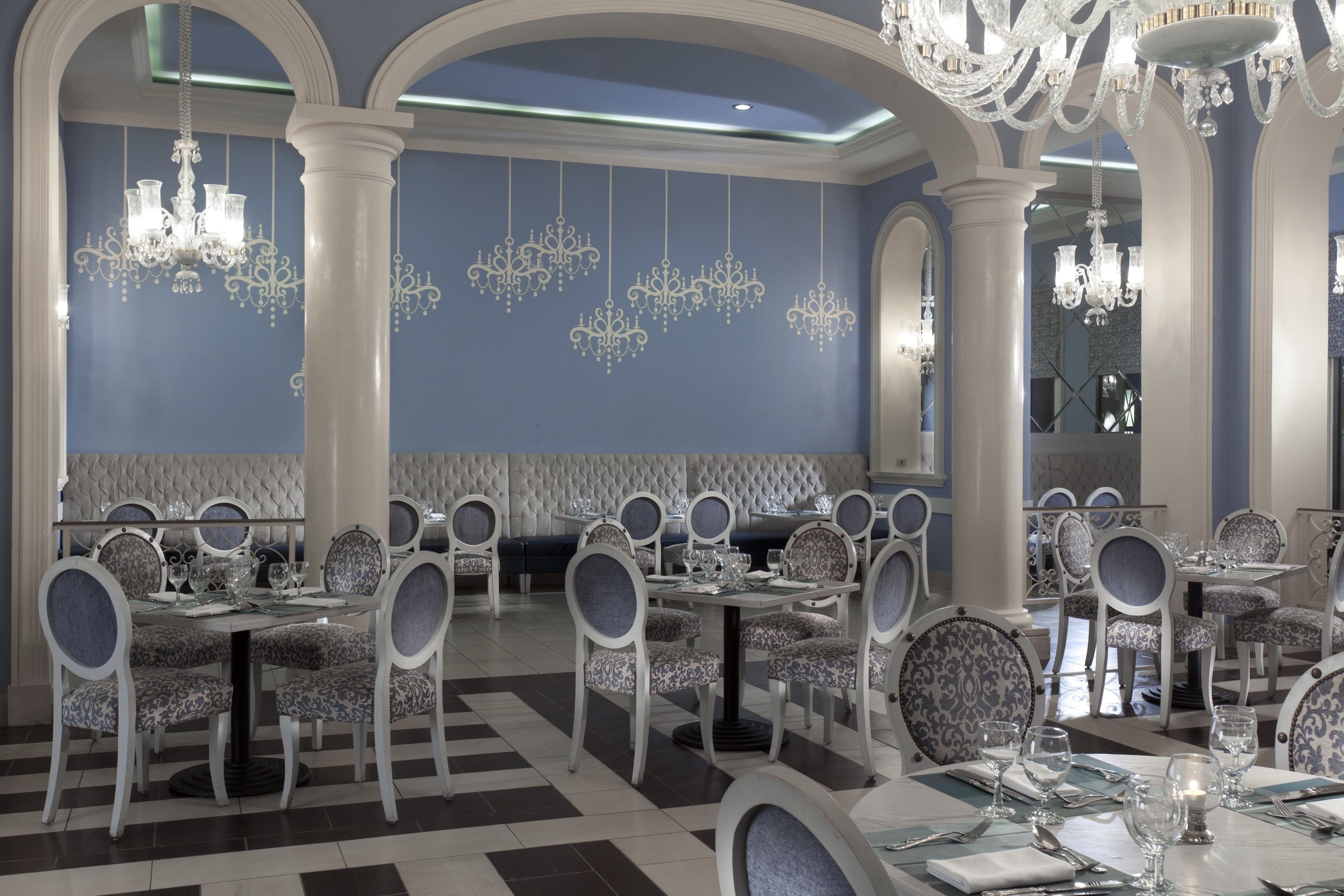 A formal dining area with multiple tables and chairs, chandeliers, and a high ceiling with decorative moldings.