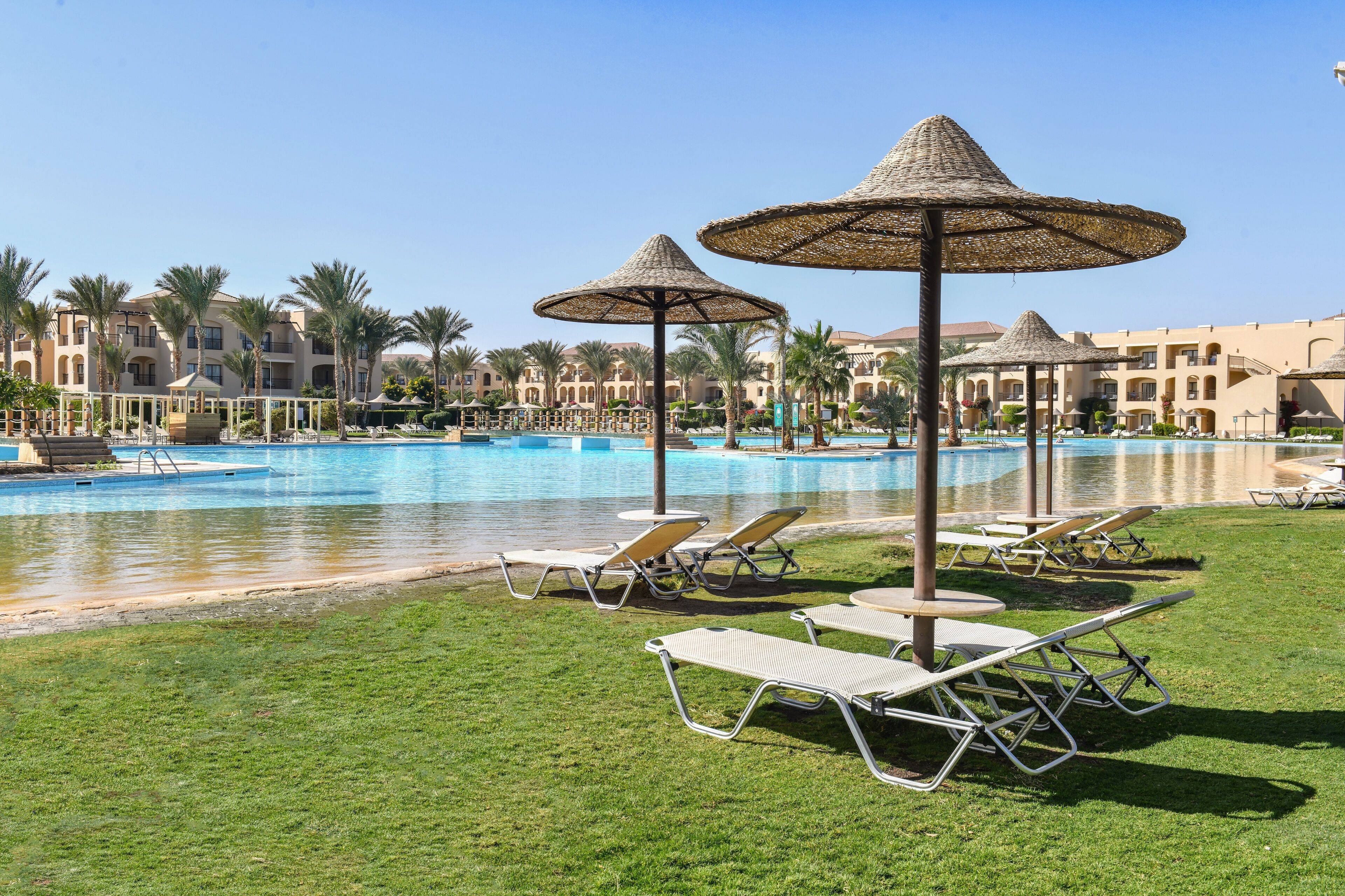 A poolside area with lounge chairs and umbrellas, surrounded by palm trees and hotel buildings.