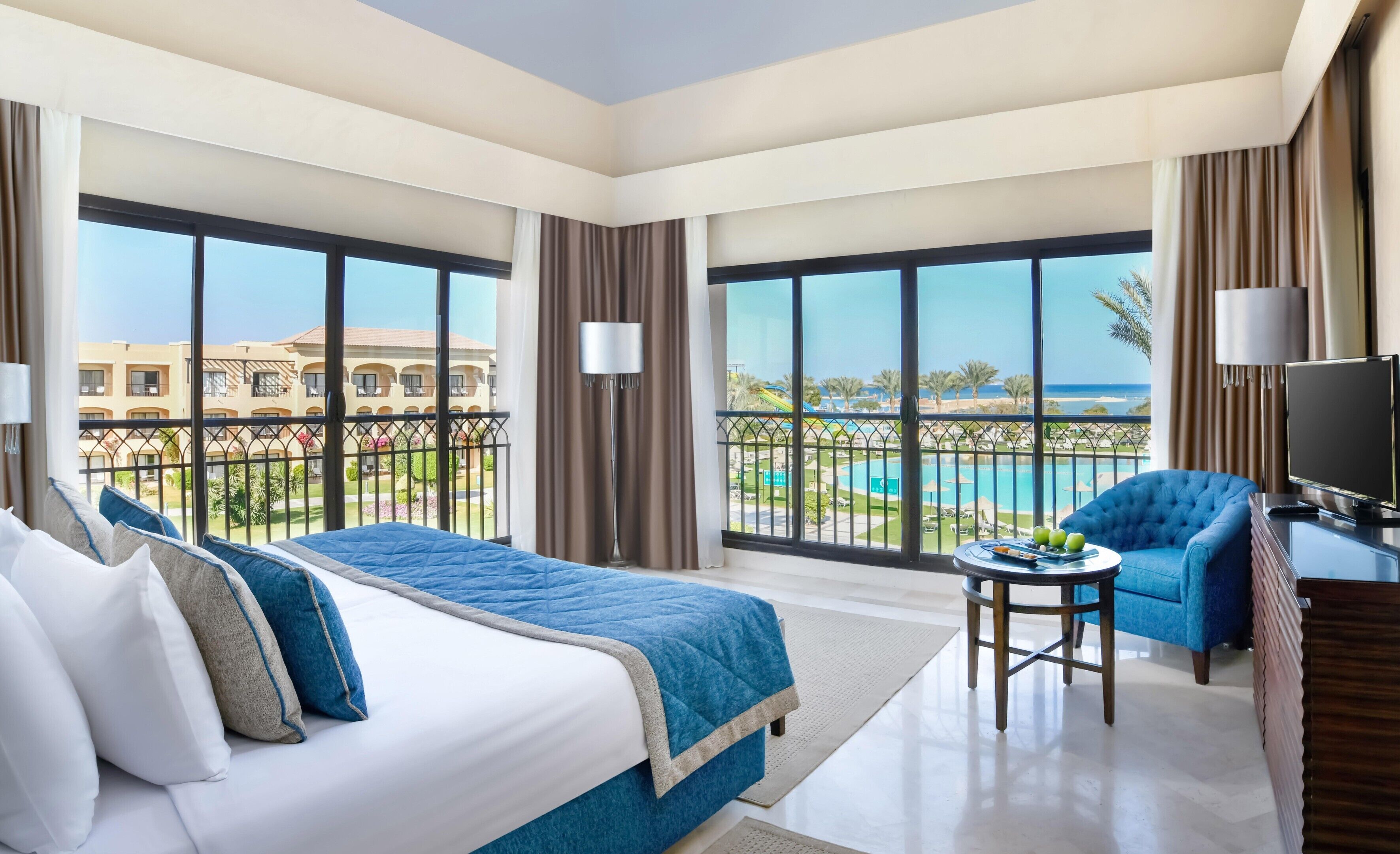 Habitación de hotel con una cama grande, un televisor, una mesita con fruta y un balcón con vista a la piscina.