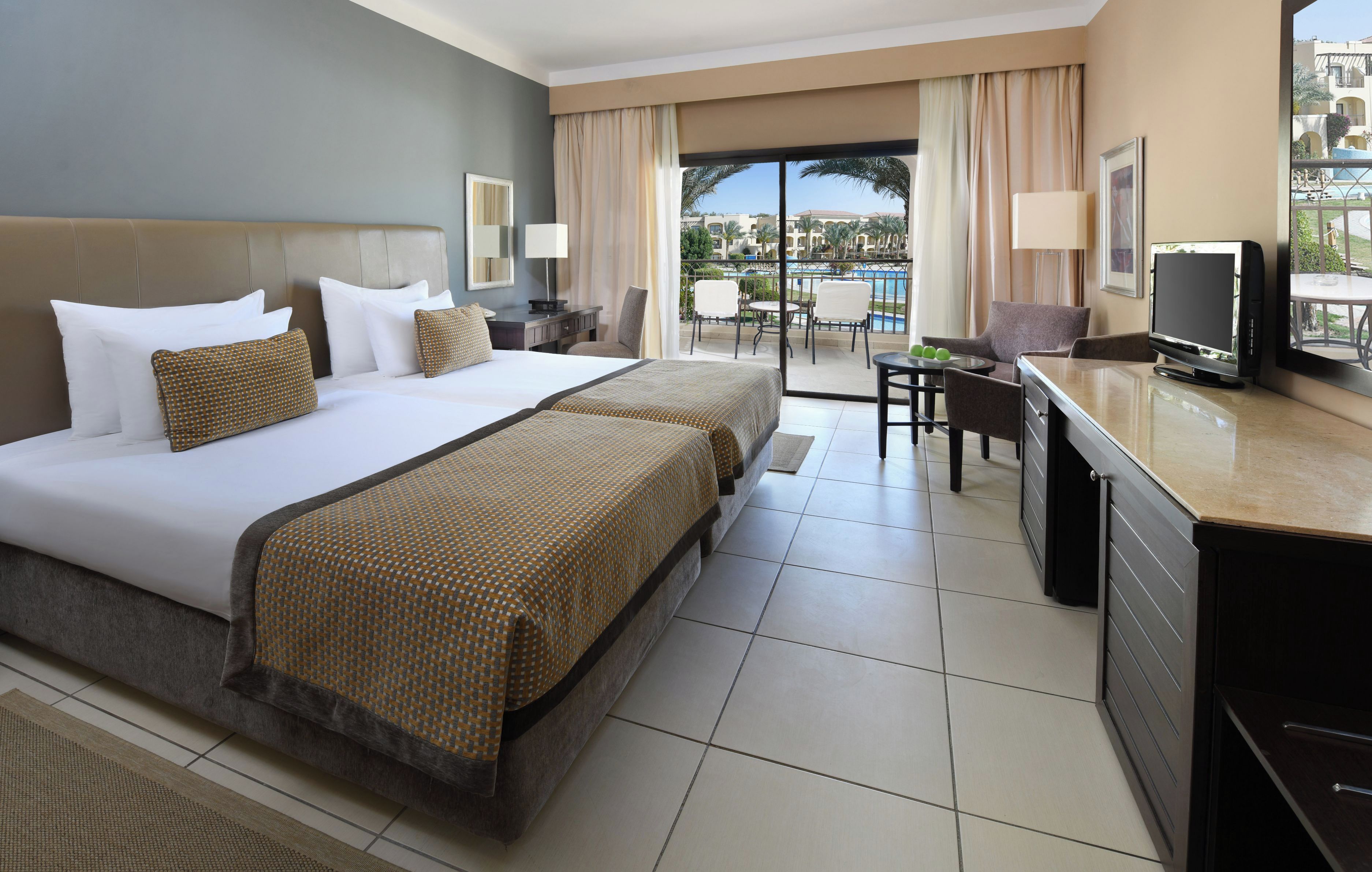 Ein Hotelzimmer mit einem großen Bett, einem Schreibtisch mit Fernseher und einem Balkon mit Blick auf den Pool und die umliegenden Gebäude.