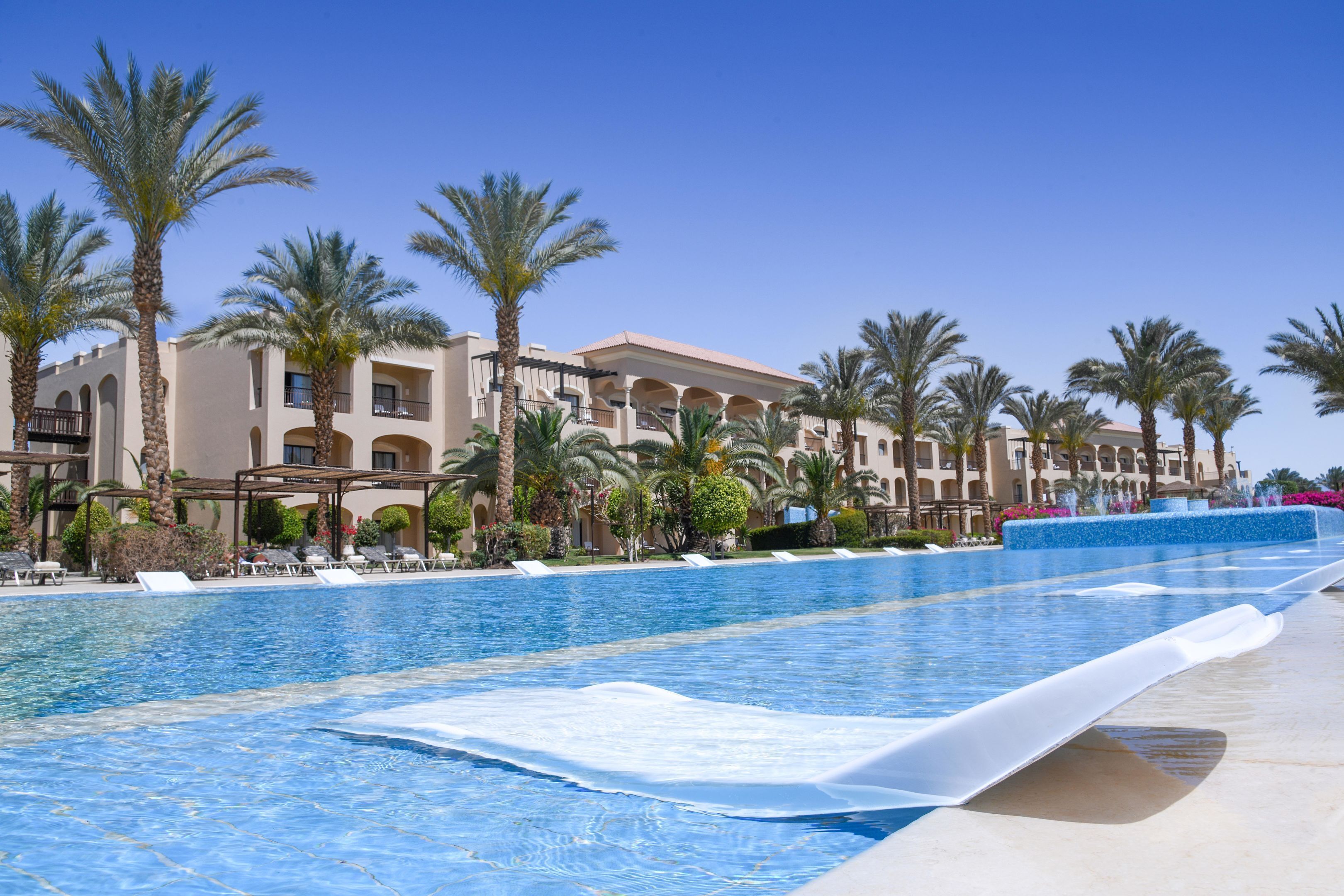 A swimming pool with lounge chairs, palm trees, and a hotel building in the background.