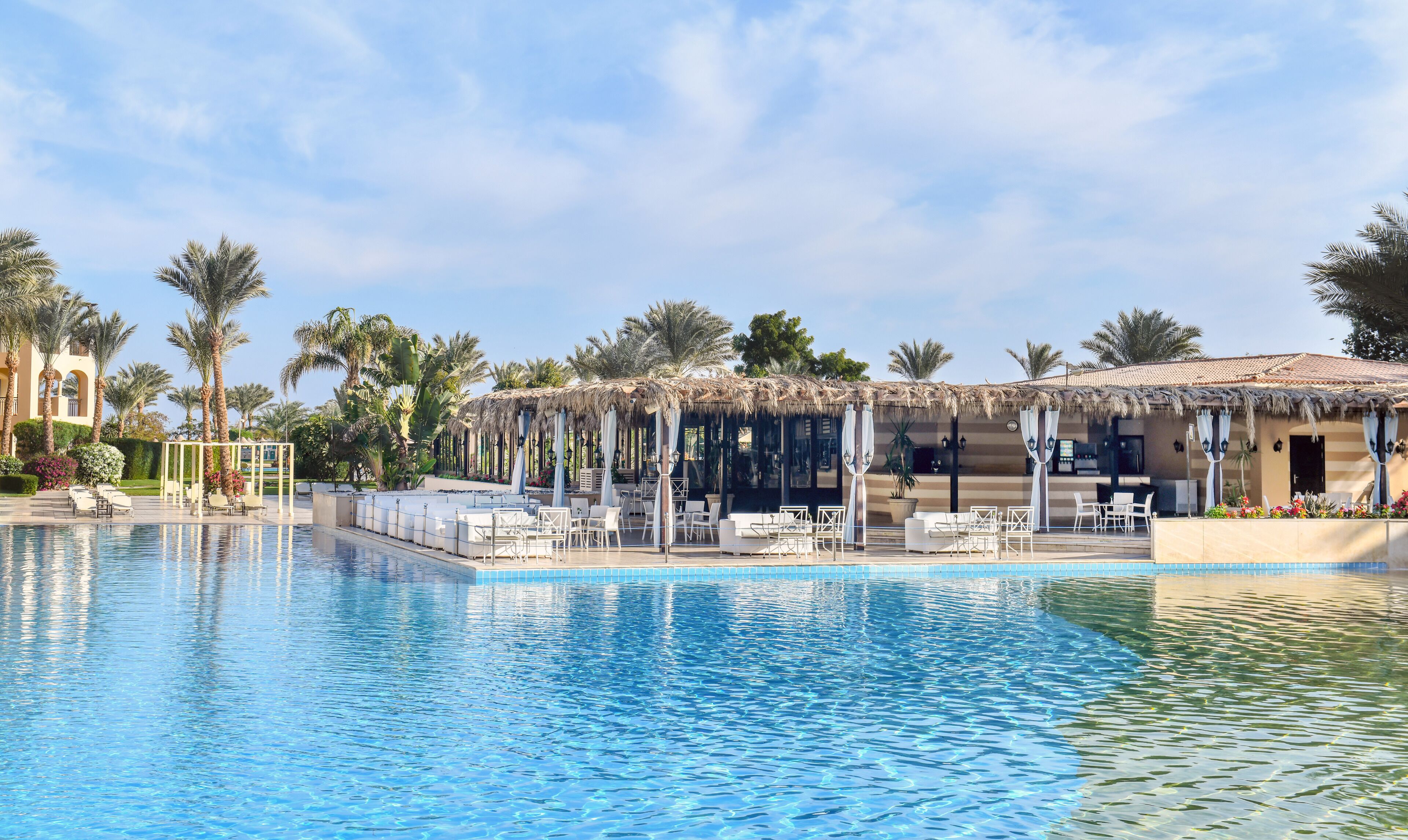 Ein Pool mit Liegestühlen und Sonnenschirmen, umgeben von Palmen und einem Gebäude mit einem Ziegeldach.