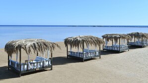 Quatre cabanes de plage aux toits de chaume, dot ées de rideaux et de coussins blancs, sont disposées sur une plage de sable fin, avec une mer d’un bleu limpide et une jetée visible au loin.