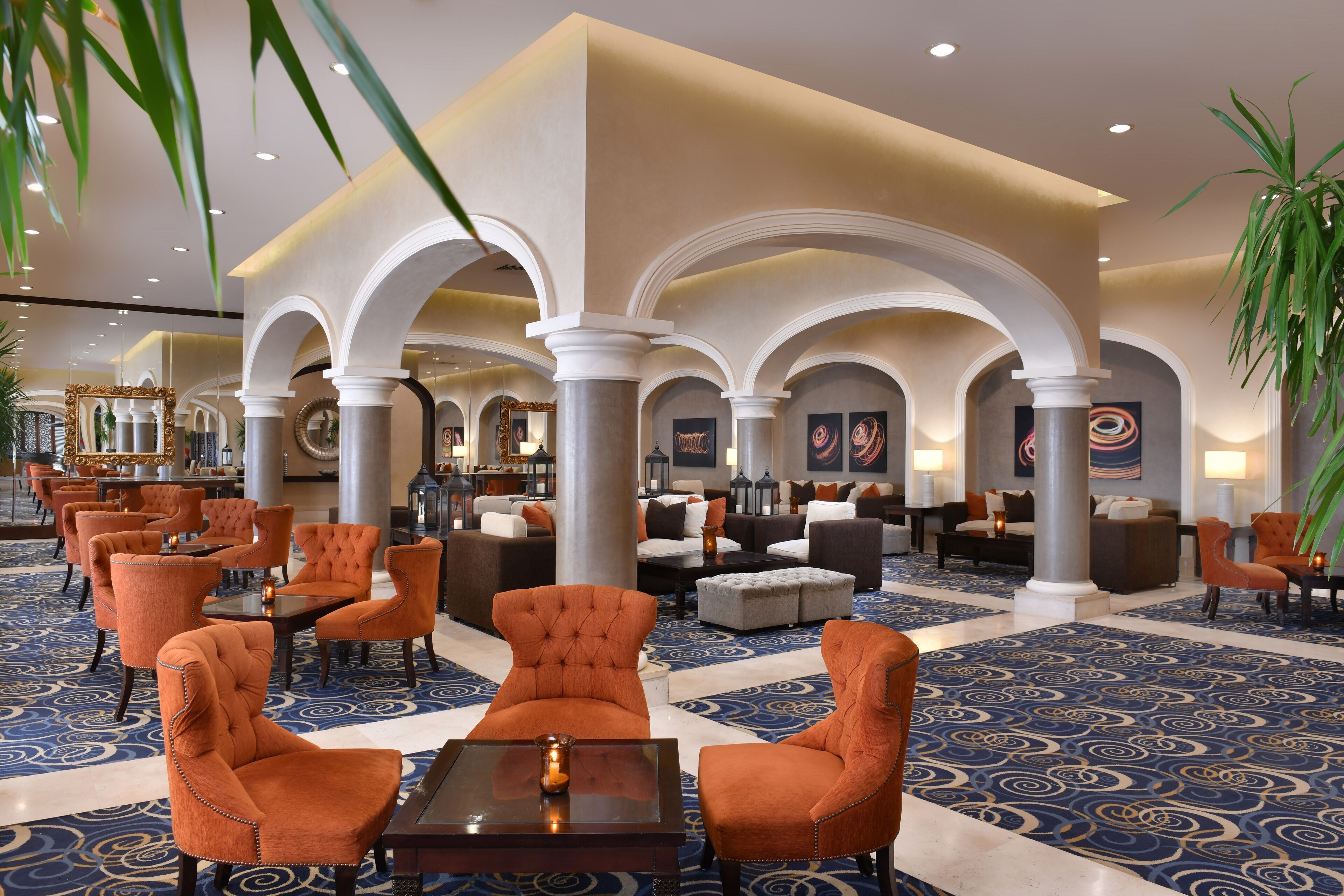 A spacious lobby with a blue patterned carpet, orange upholstered chairs, and a central seating area with sofas and tables.