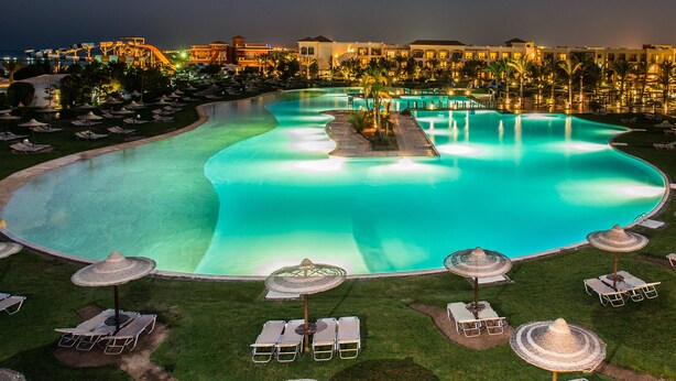 Une grande piscine incurvée, entourée de chaises longues et de parasols, avec un bâtiment de complexe hôtelier en arrière-plan.