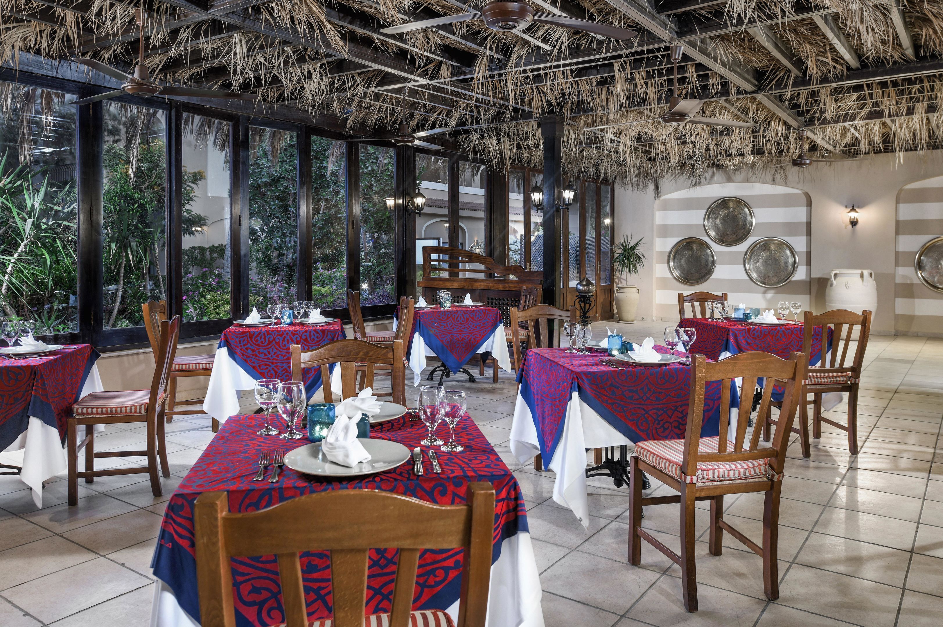 A restaurant with tables set for dining, a thatched roof, and large windows.