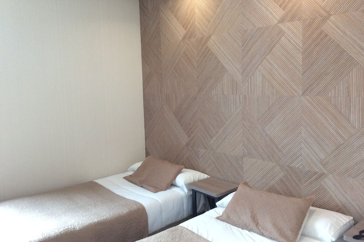 A hotel room with two single beds, a small table, and a geometric patterned wall.