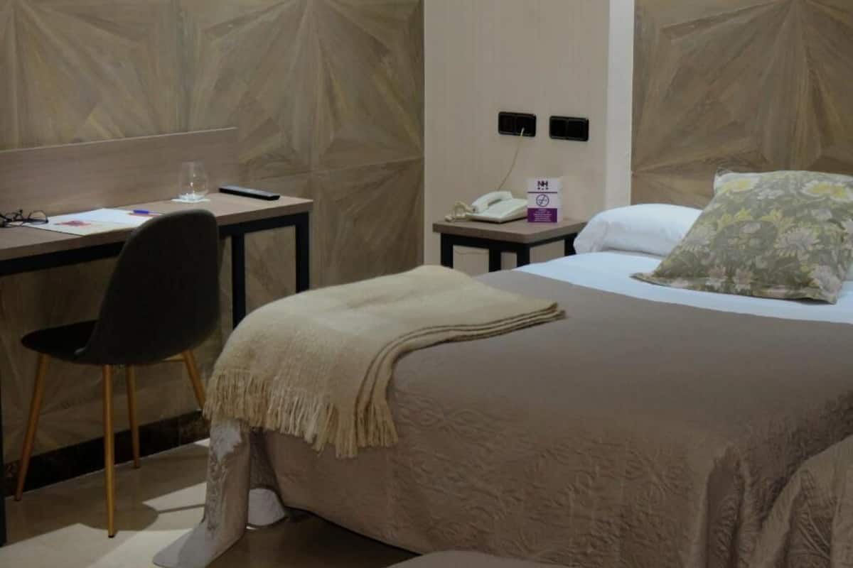 A hotel room with a bed, a desk, a chair, a phone, and a wall with a geometric pattern.