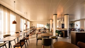 Ein geräumiger Hotel-Speiseraum mit mehreren Tischen und Stühlen, einer zentralen Bar und großen Fenstern für natürliches Licht.