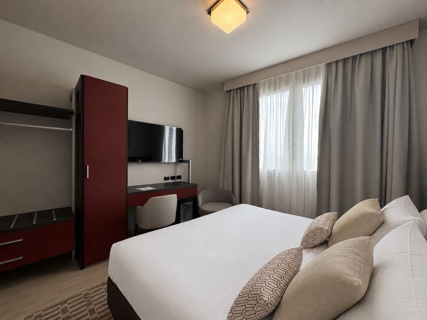 Une chambre d’hôtel avec un lit, un bureau avec une télévision, une armoire et une fenêtre avec des rideaux.