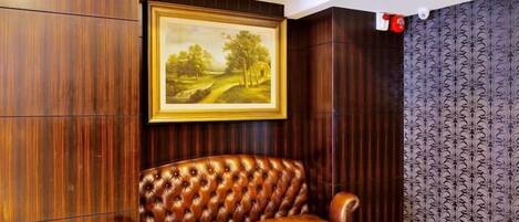 A room with a brown leather sofa, a framed landscape painting, wooden paneling, and patterned wallpaper.