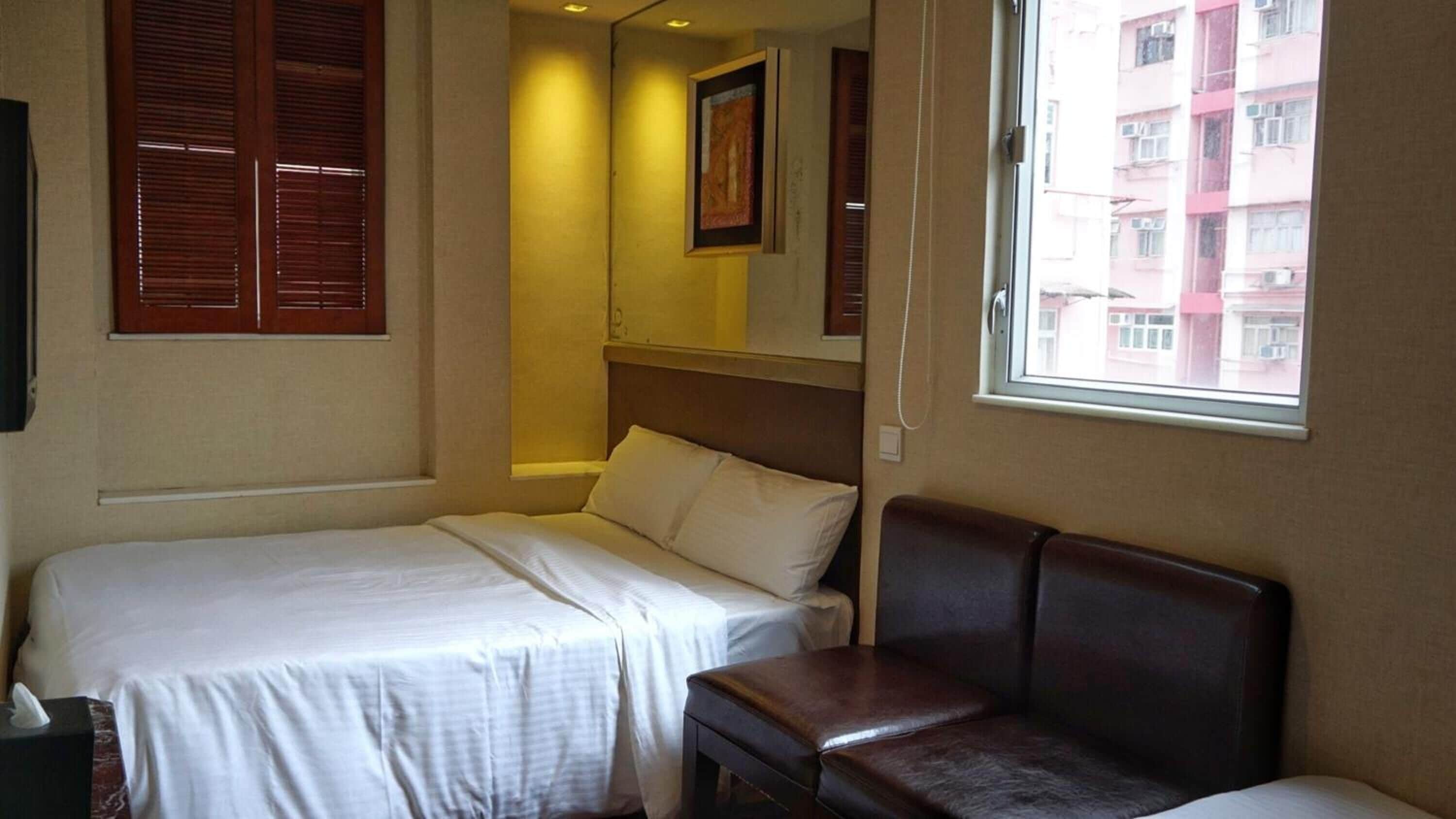 A hotel room with two beds, a bench, a window with a view of buildings, and a framed picture on the wall.
