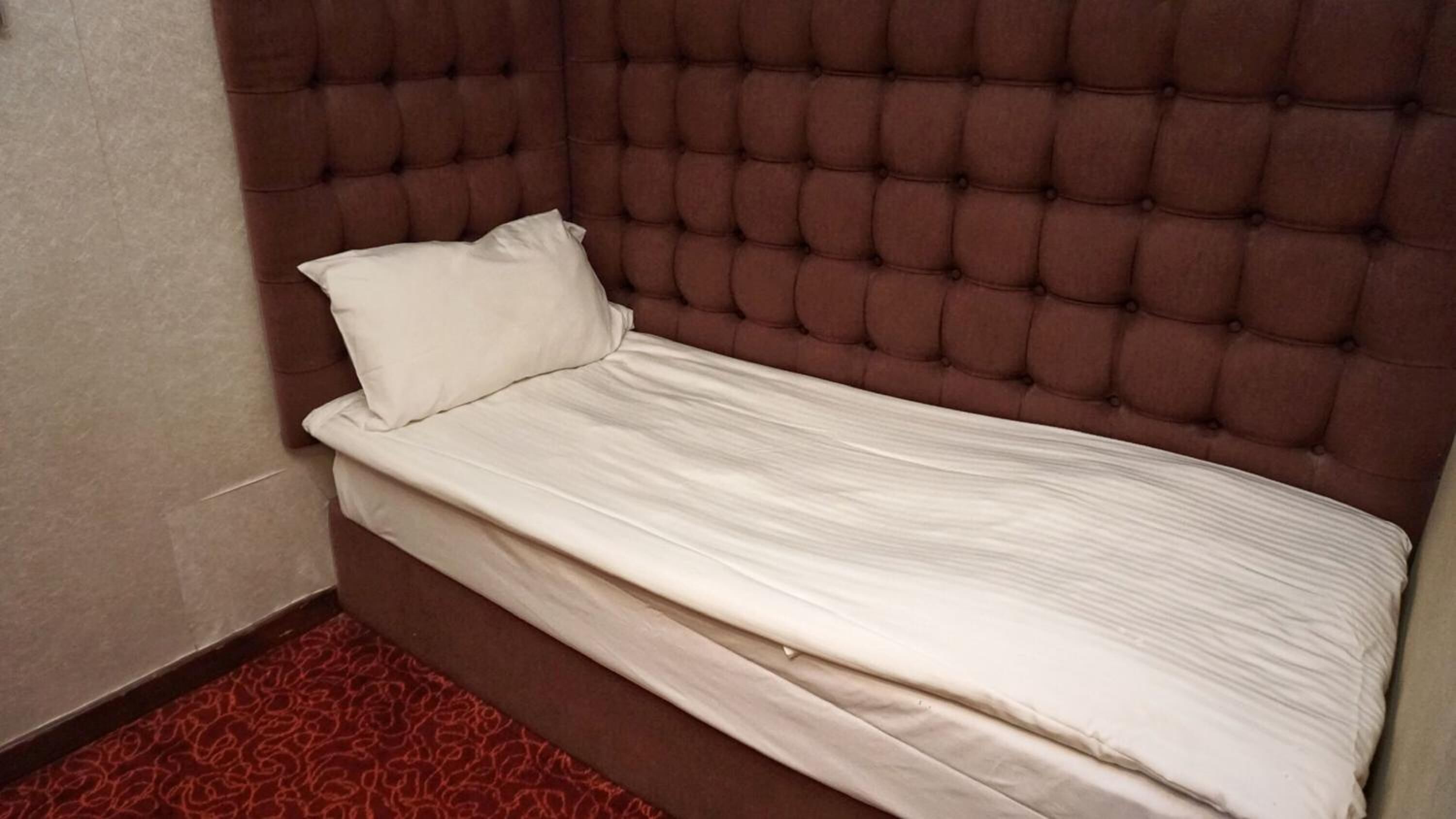 A small, single-bed room with a tufted headboard and a red patterned carpet.