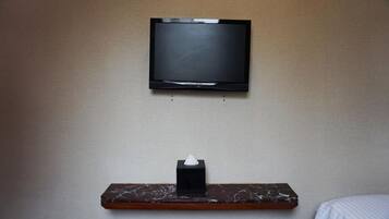 A television mounted on a wall, a tissue box on a wooden shelf below, and a bed with a white sheet.