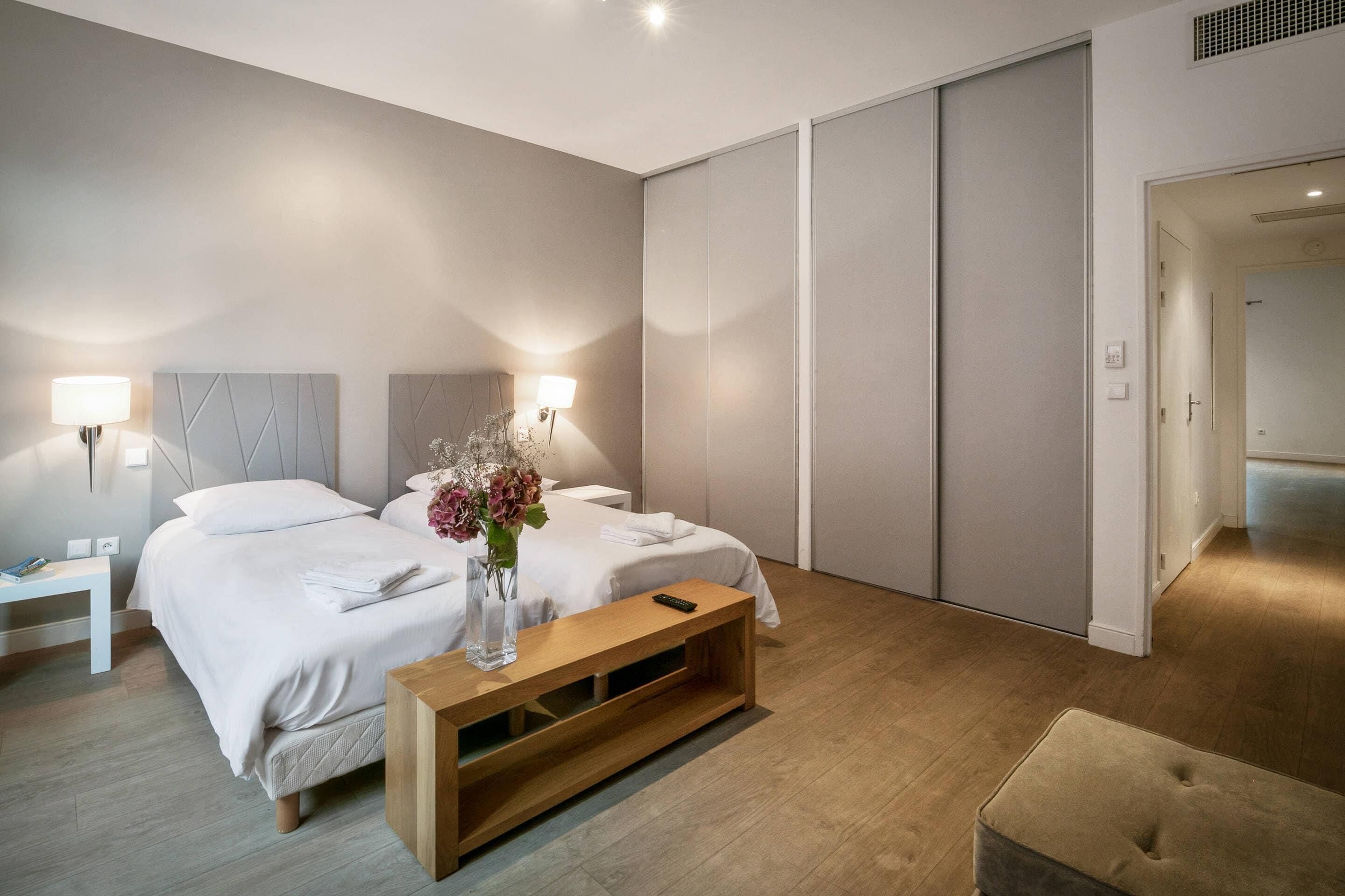A modern hotel room with a large bed, bedside lamps, a wooden bench, and a small table with a vase.
