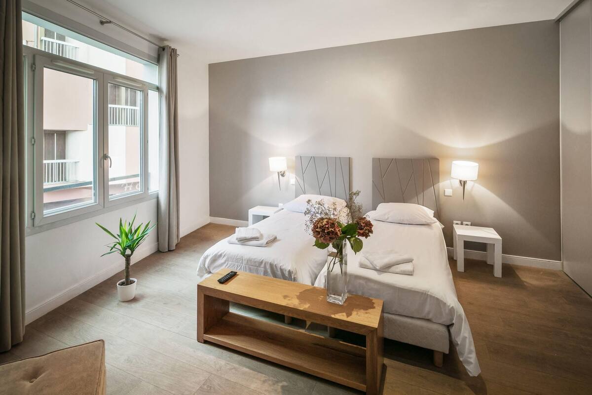 Une chambre d’hôtel moderne avec deux lits, un banc en bois, une table de chevet, un vase avec des fleurs et une vue sur le balcon extérieur.