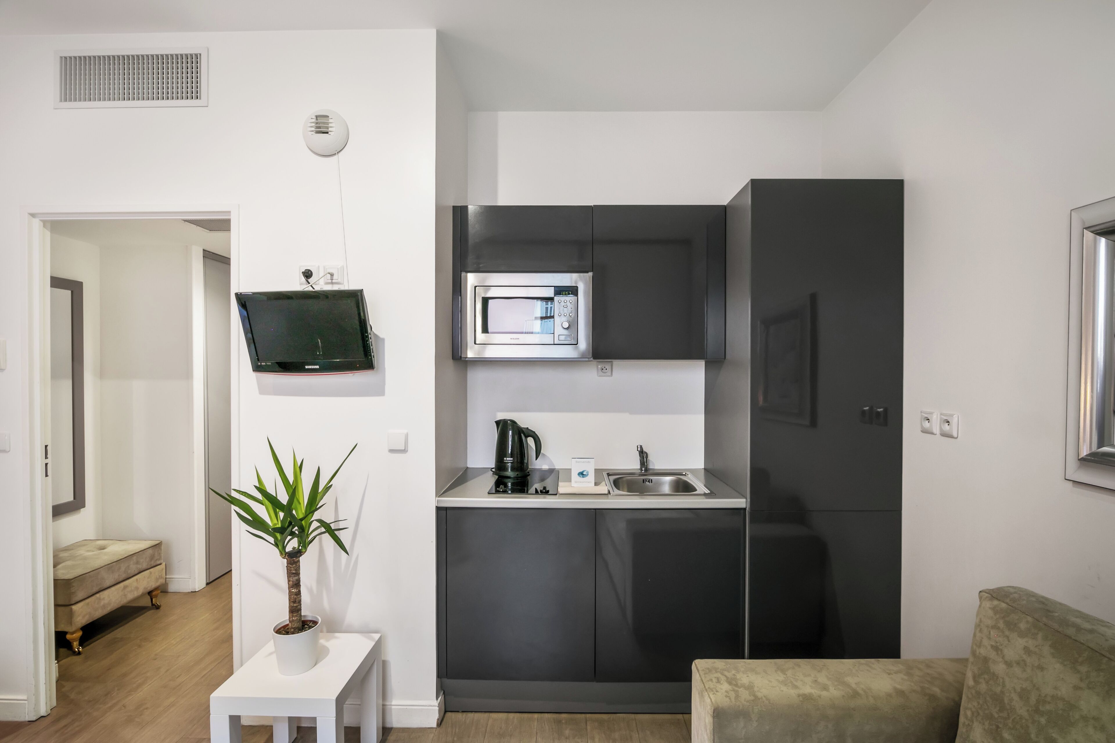A compact kitchen with a microwave, coffee maker, and sink. There is a small white table with a plant and a beige sofa in the living area.