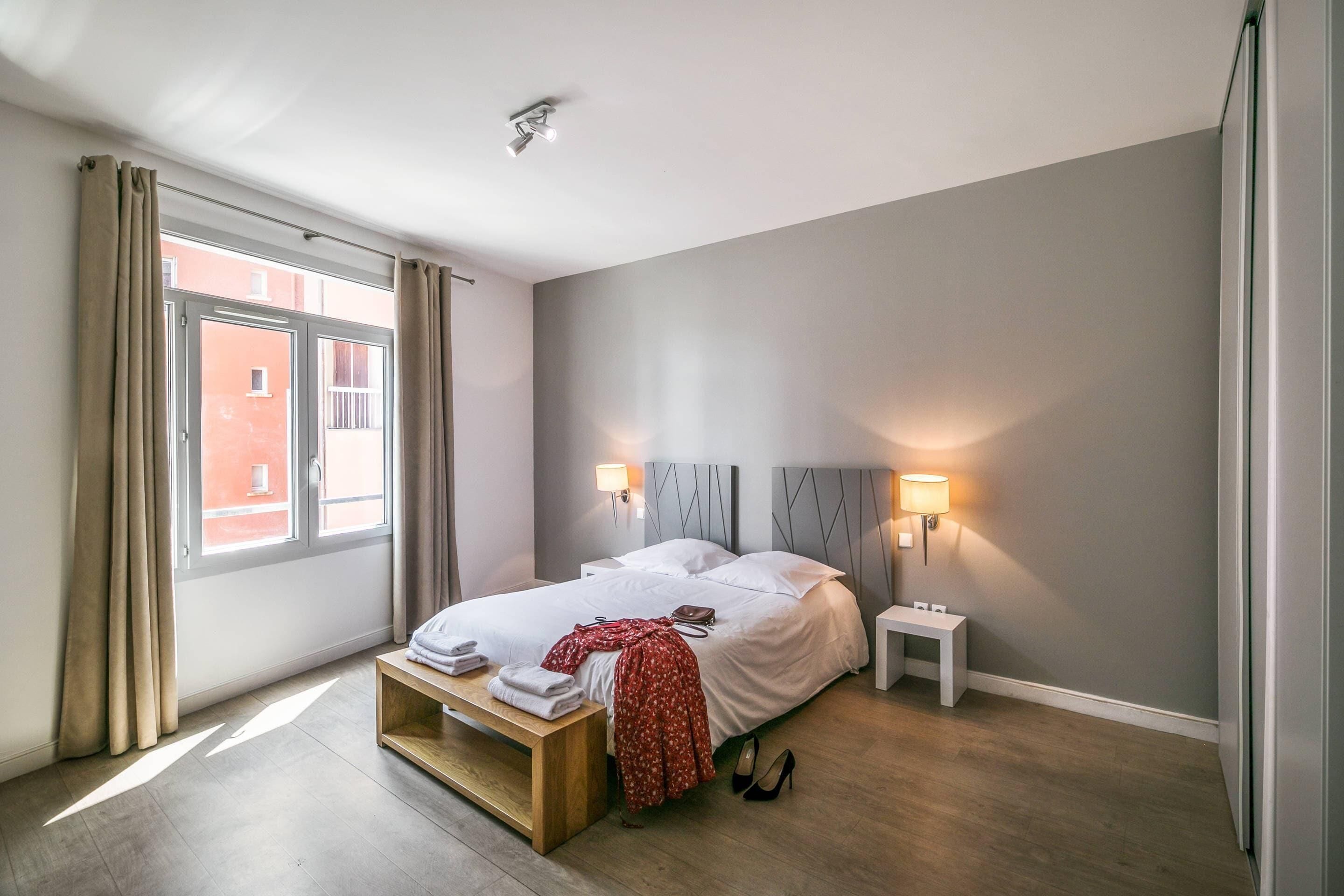 A modern bedroom with a bed, bedside tables, a bench, and a view of a building through the window.