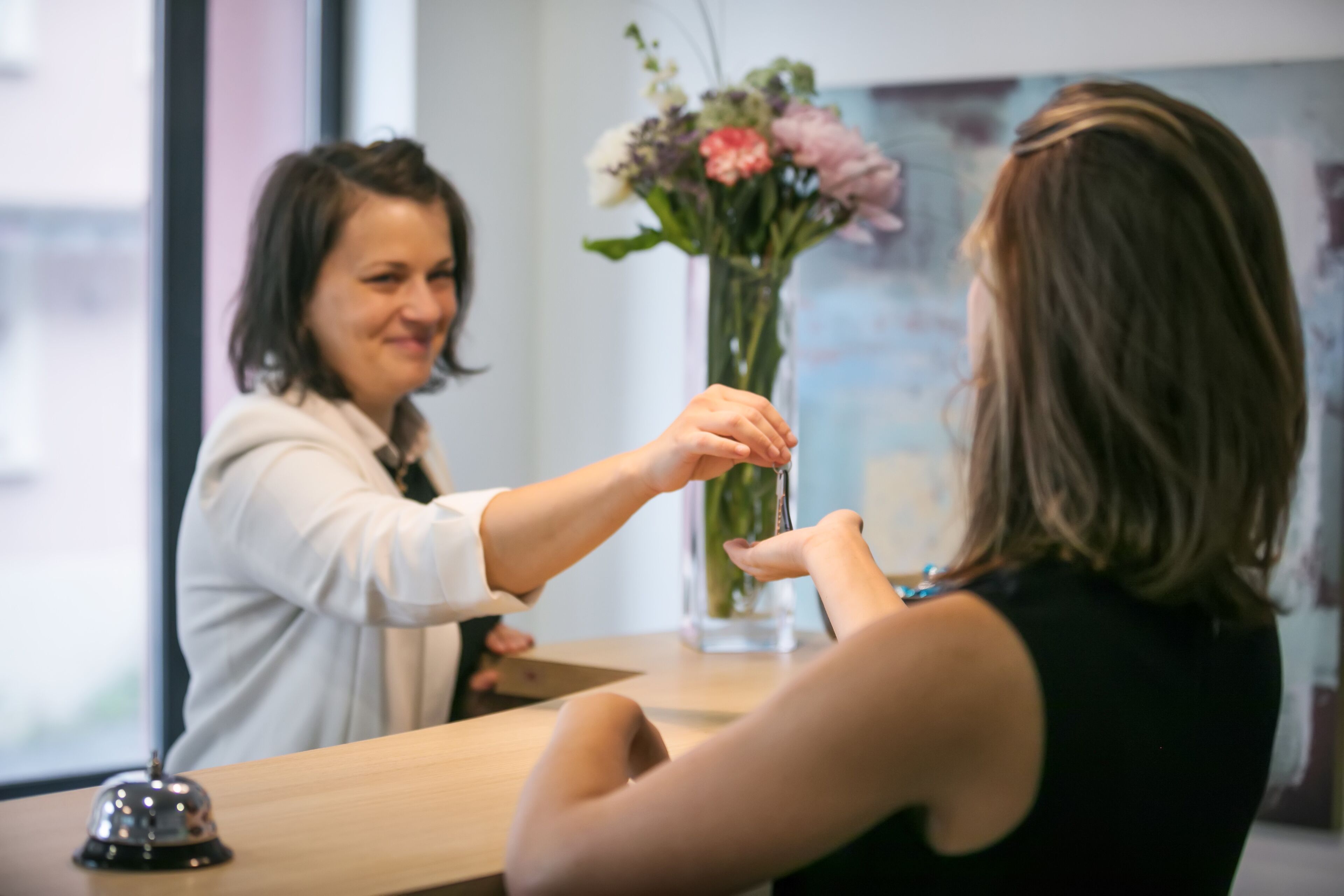 A reception desk with a bell, a vase with flowers, and a window in the background.
