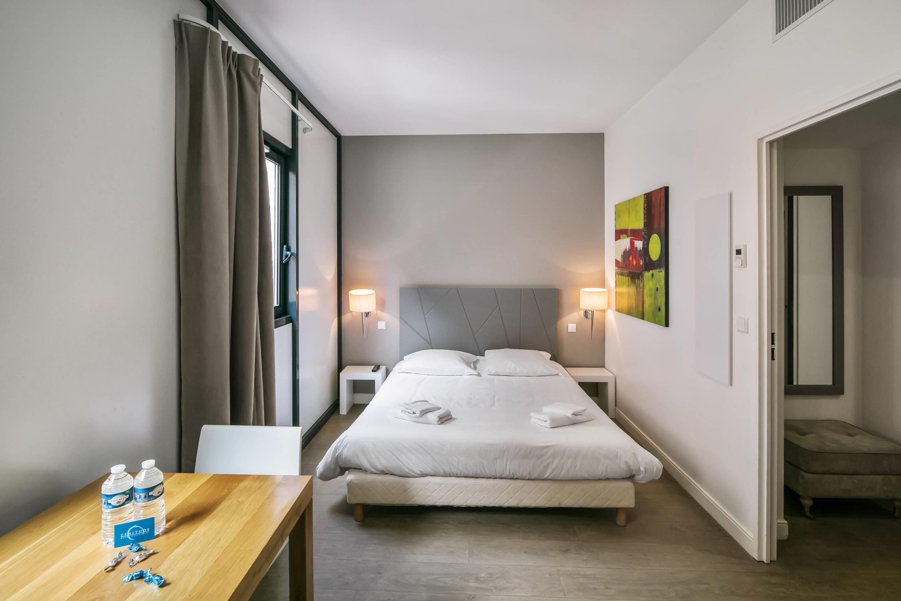 A hotel room with a bed, bedside tables, a desk with water bottles, and a painting on the wall.