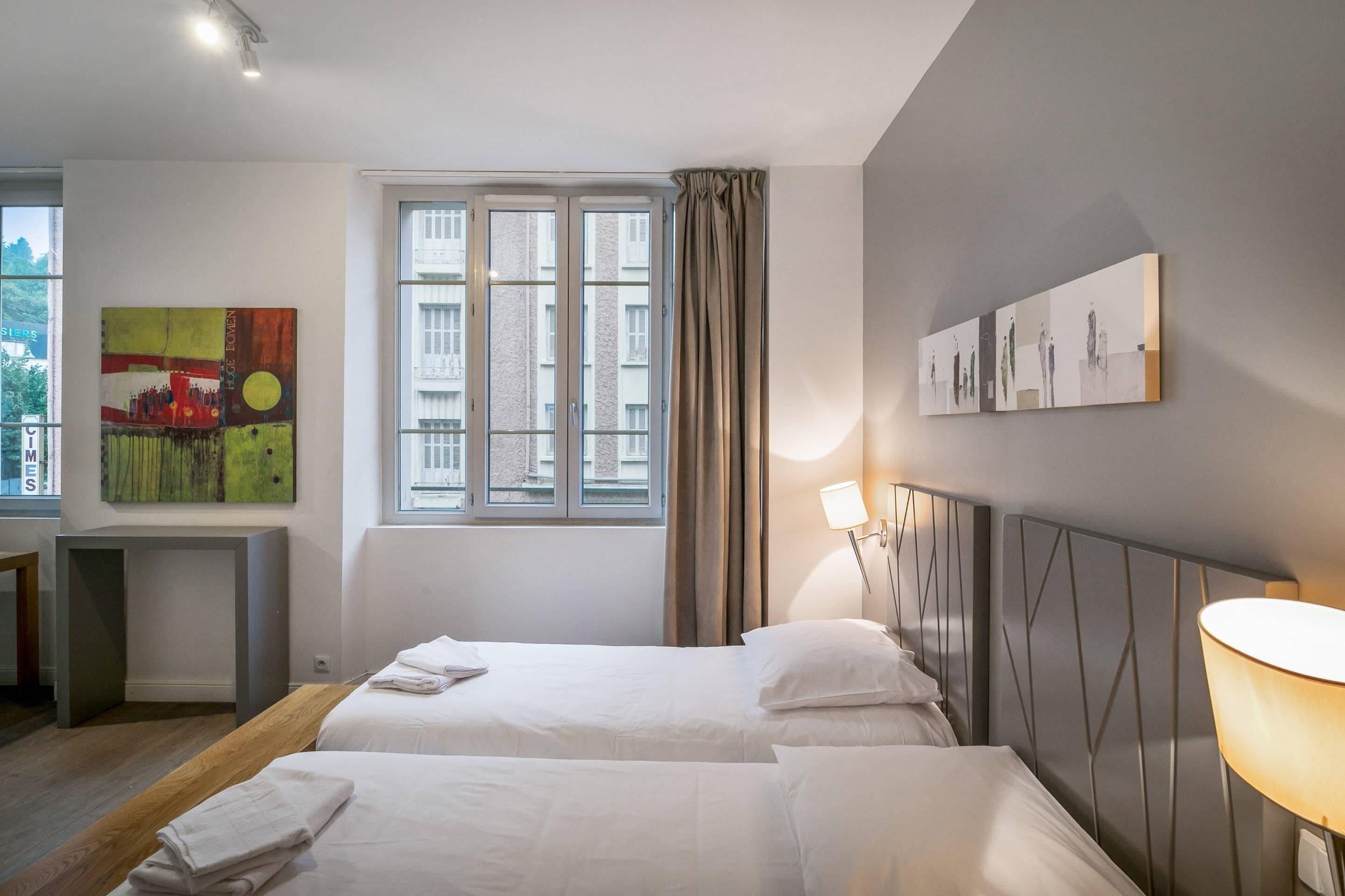 Une chambre d’hôtel avec deux lits, une fenêtre avec des rideaux, un bureau et des œuvres d’art accrochées aux murs.