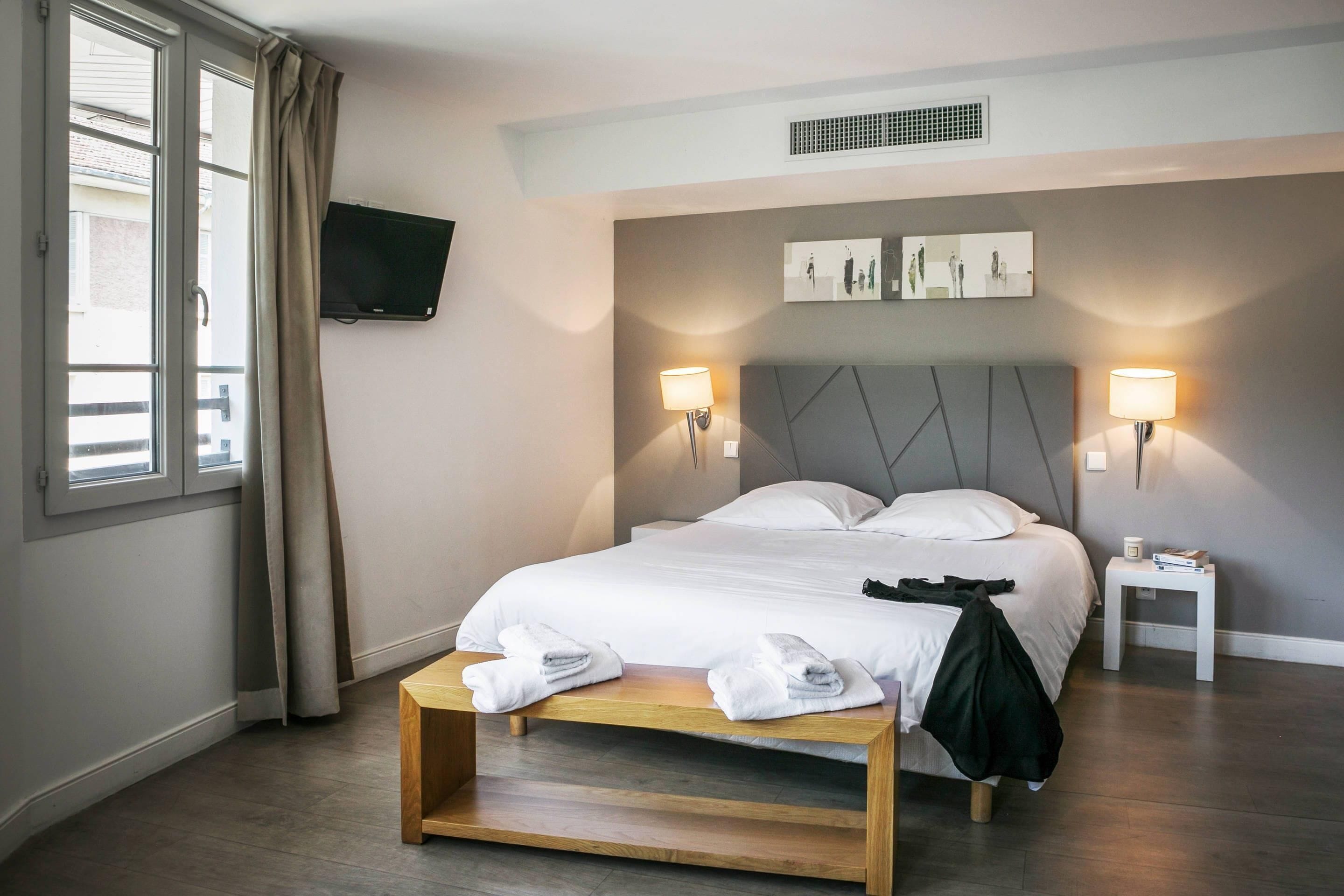 A hotel room with a bed, bedside lamps, a TV mounted on the wall, and a window with curtains.